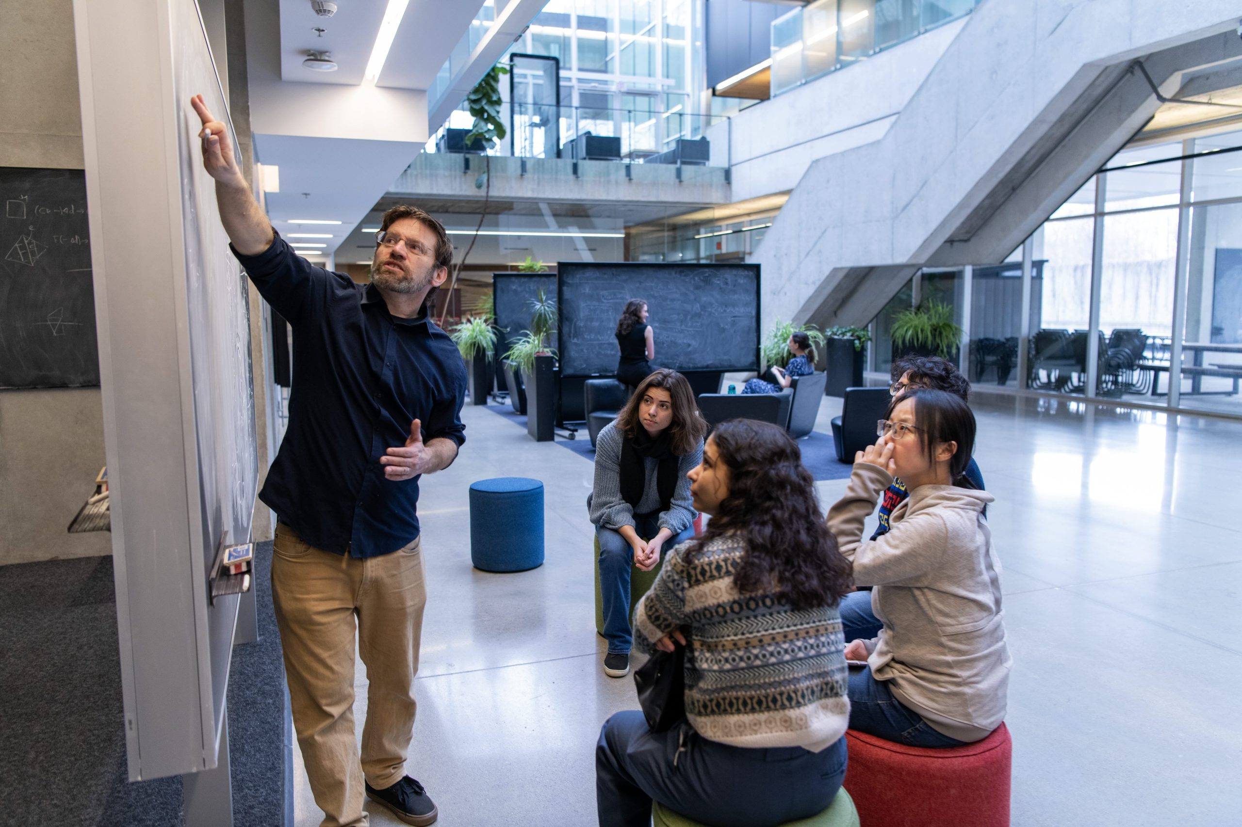 a group of people sitting while another person stands and points to something on a blackboard